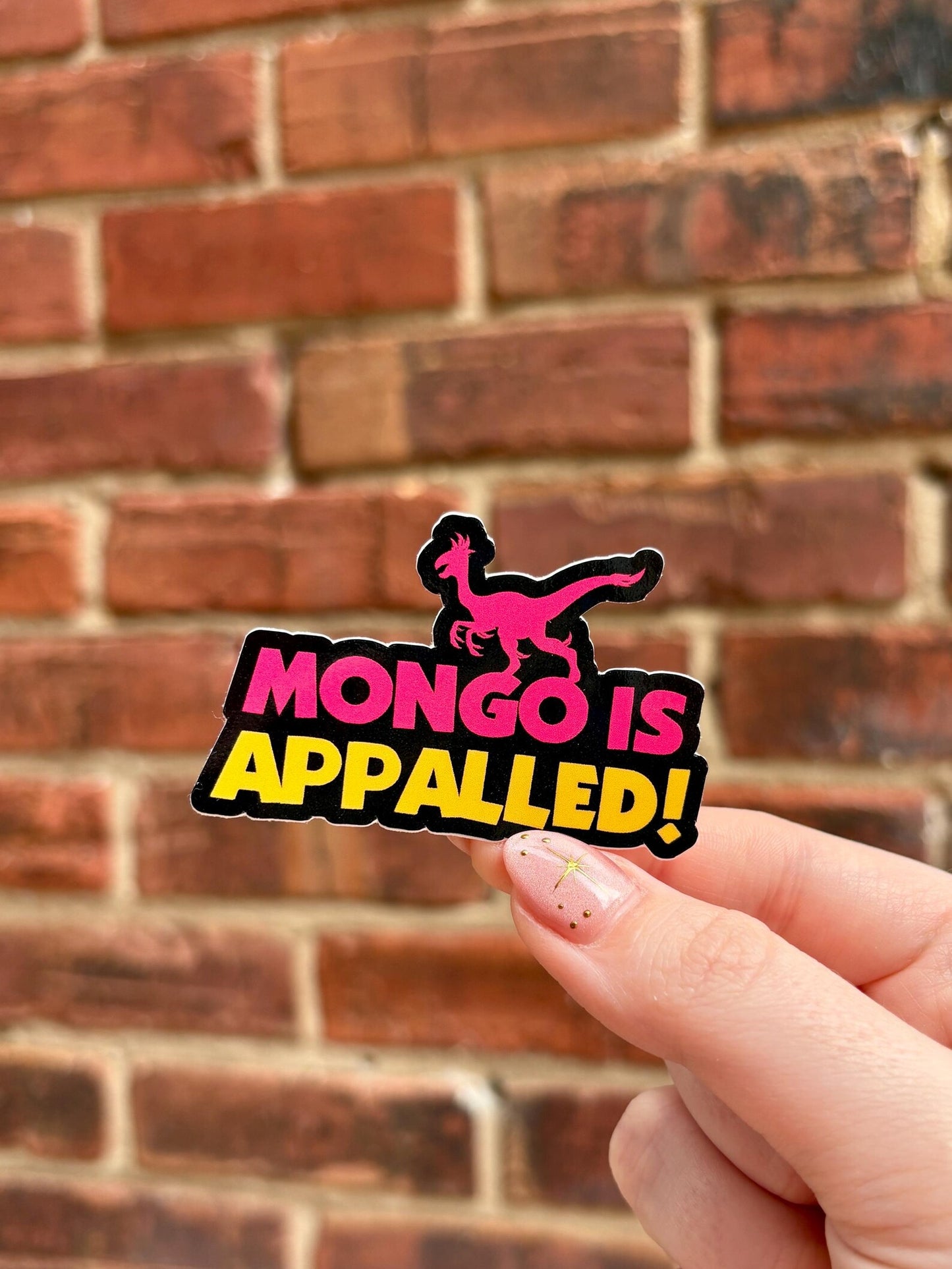 Hand holding a sticker with 'Mongo is Appalled!' text against a brick wall background
