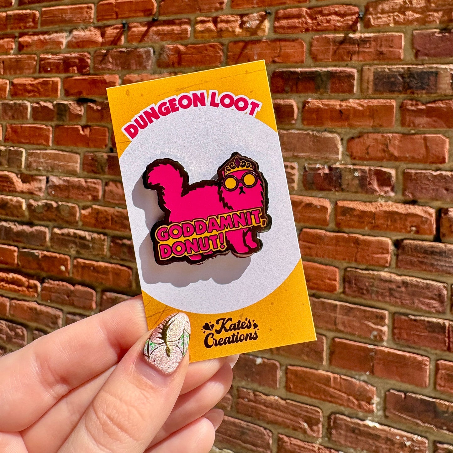 Pin with 'Dungeon Loot' and 'Godammit Donut!' design held against a brick wall.