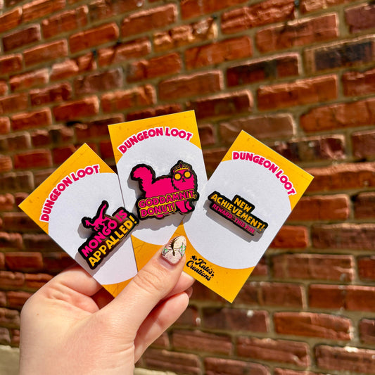 Hand holding a set of 'Dungeon Loot' pins against a brick wall.
