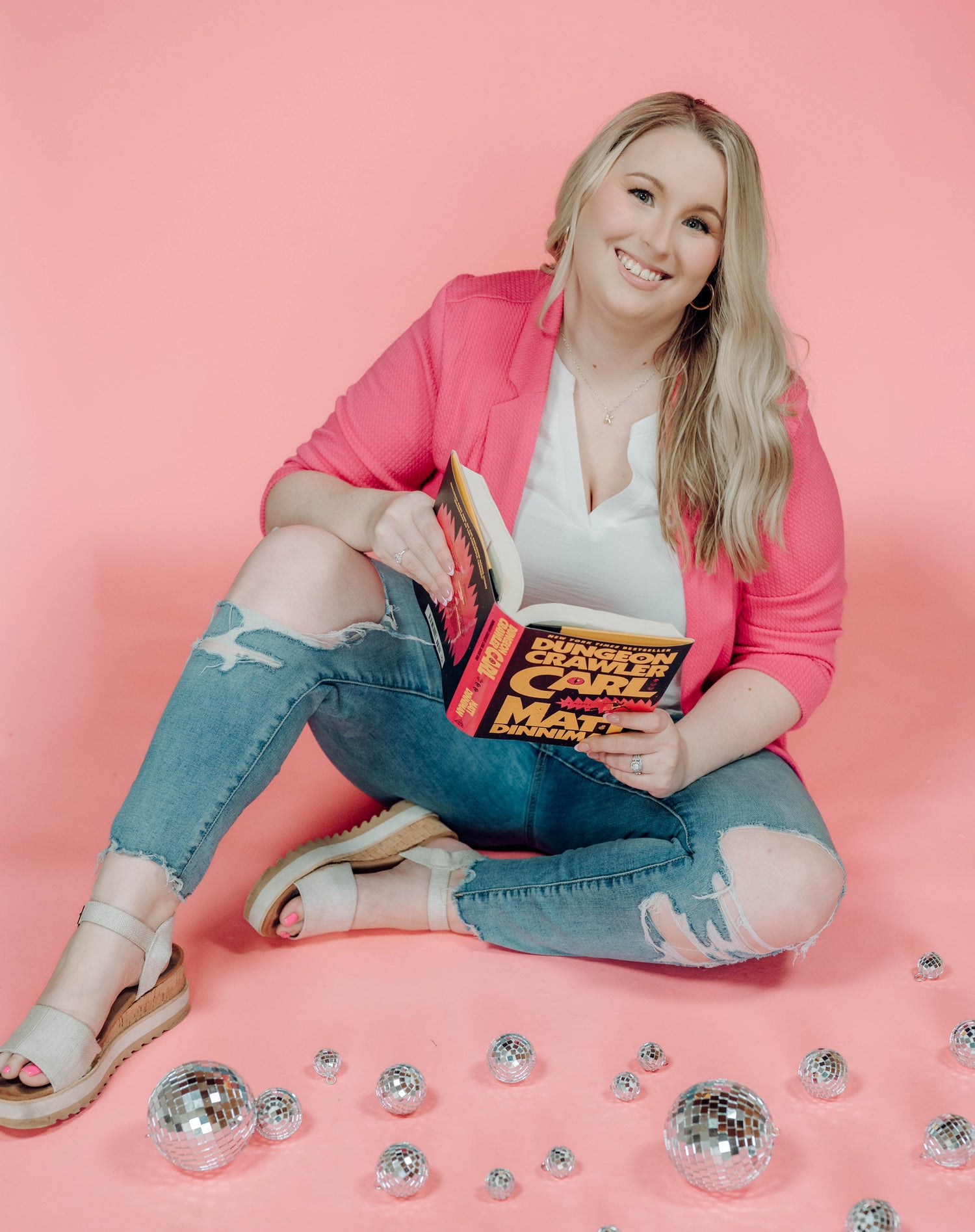 Woman sitting on a pink background holding a book, surrounded by decorative items.