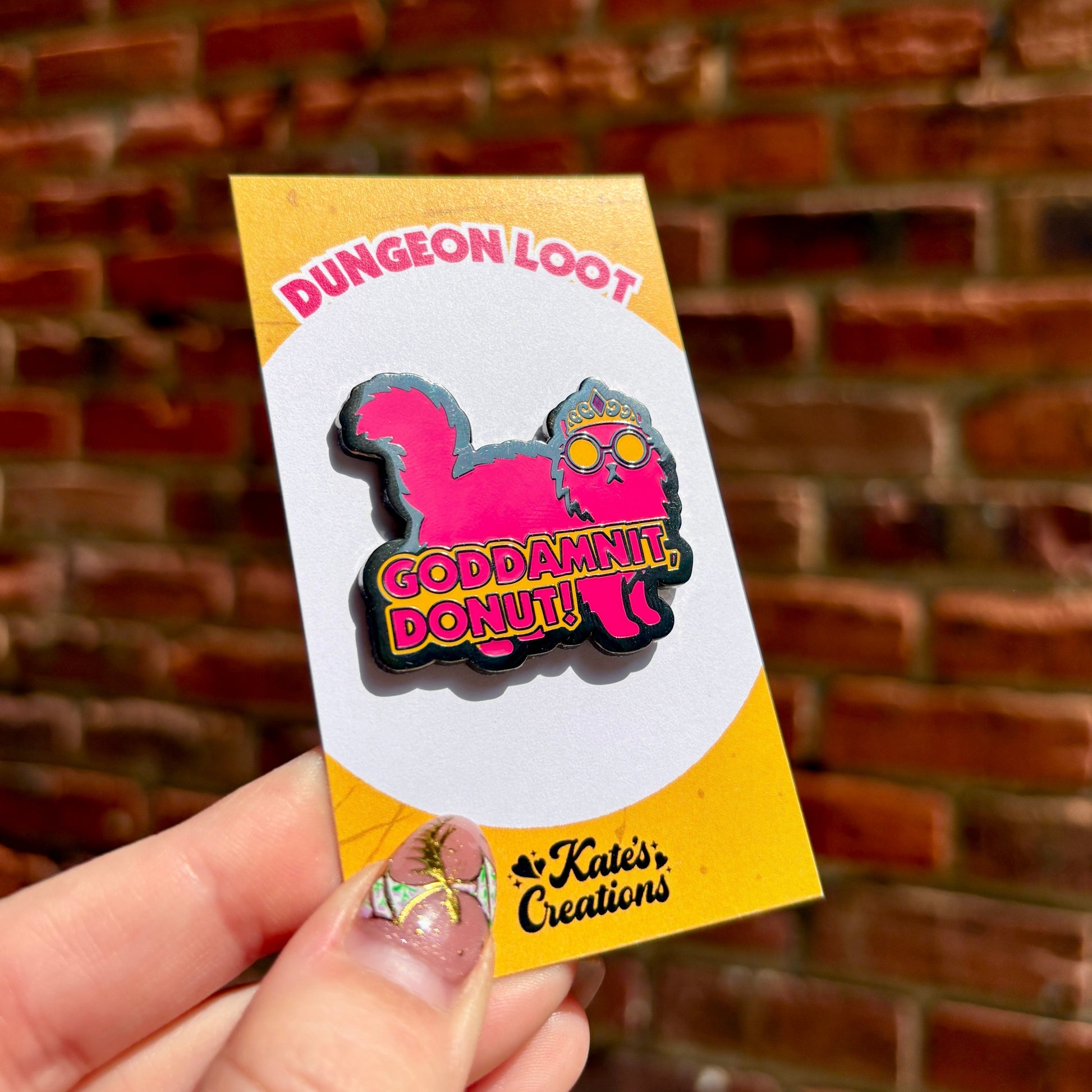 Hand holding a 'Dungeon Loot' sticker with a pink donut design against a brick wall.