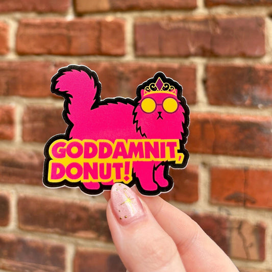 Sticker with a pink cat design and text 'GODDAMNIT, DONUT!' held against a brick wall.