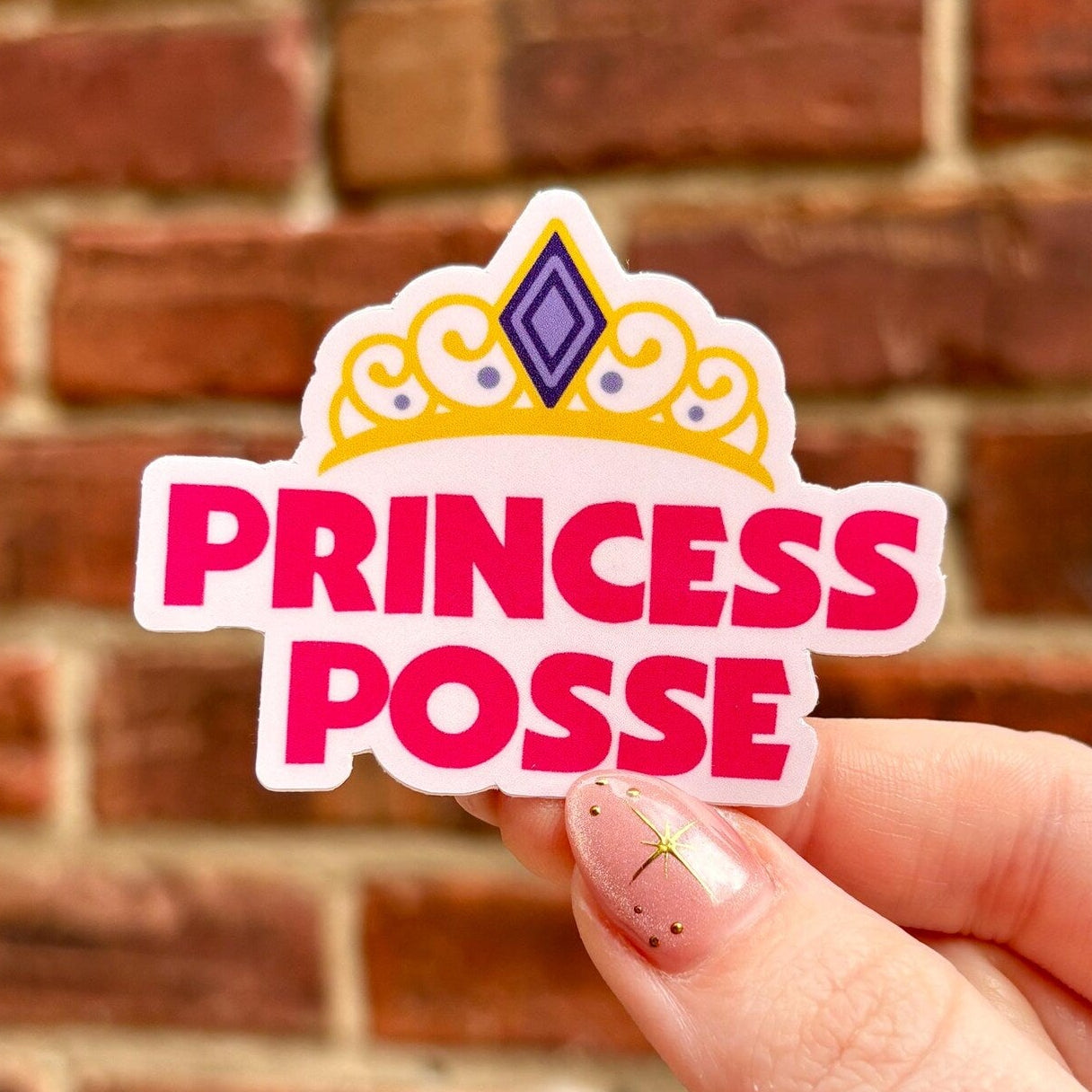 Hand holding a 'Princess Posse' sticker against a brick wall